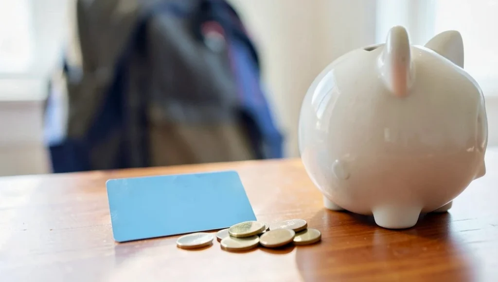 Blank debit card, a small pile of coins, and a piggy bank on a desk with a school backpack blurred in the background